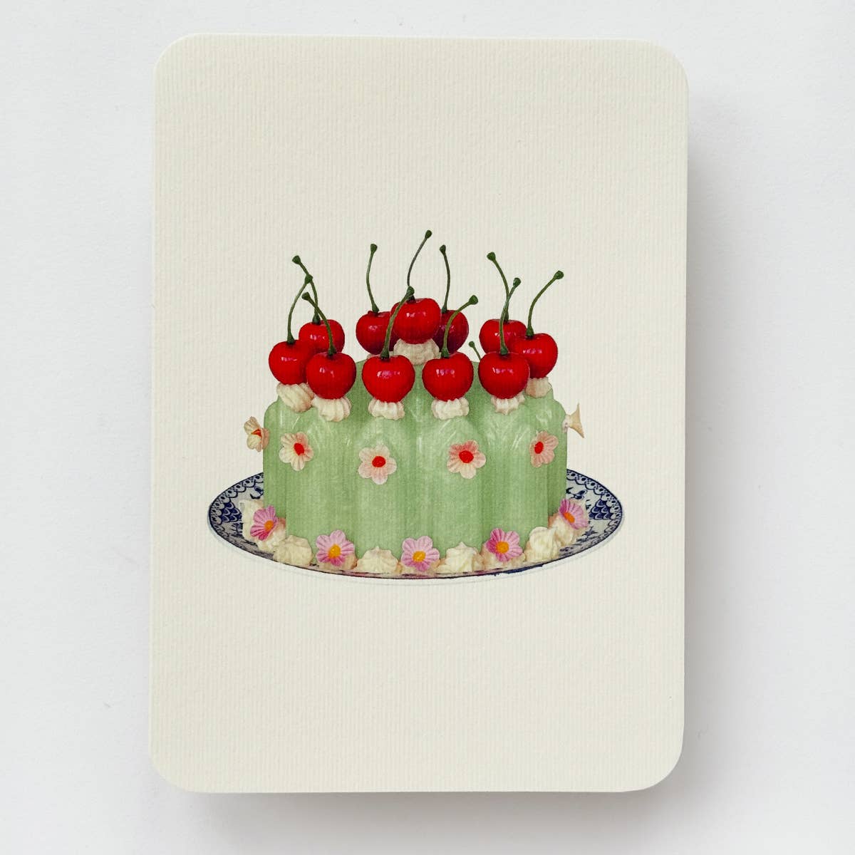 Greeting card with cream background and image of a green Blancmange dessert with red stemmed cherries and dollops of white cream on blue and white plate. Envelope included.