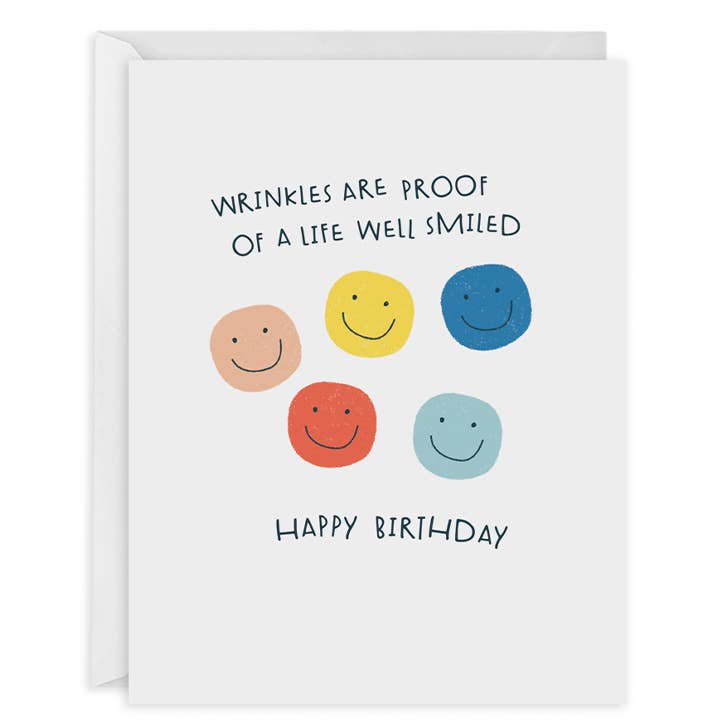 Greeting card with white background and images of smiley faces in peach, yellow, blue, red and aqua and black text says, "Wrinkles are proof of a life well smiled, Happy birthday". White envelope included, 