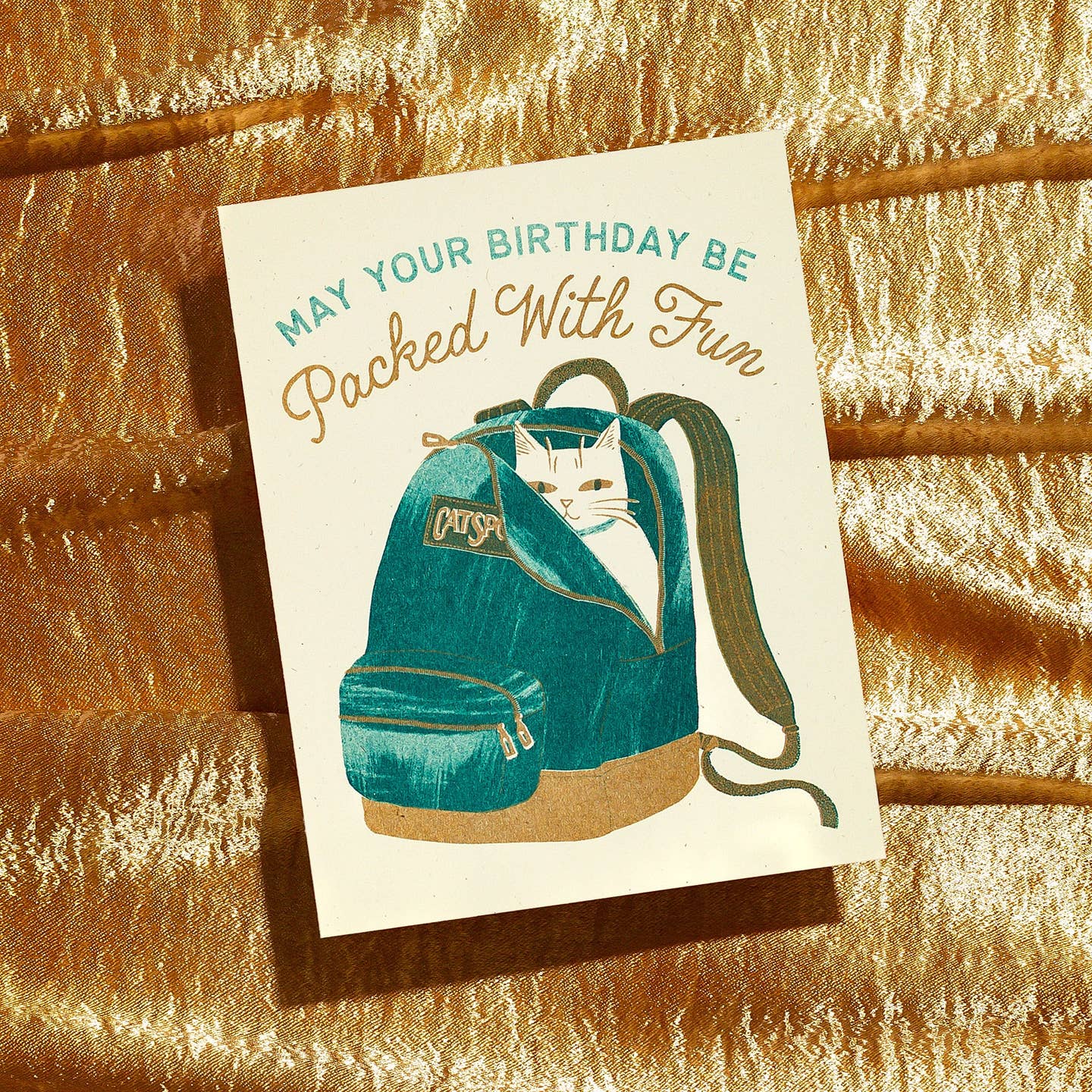 Greeting card with  cream background and image of a white cat in a teal backpack with teal text says, "May your birthday be" and tan text says, "Packed with fun". Envelope included.