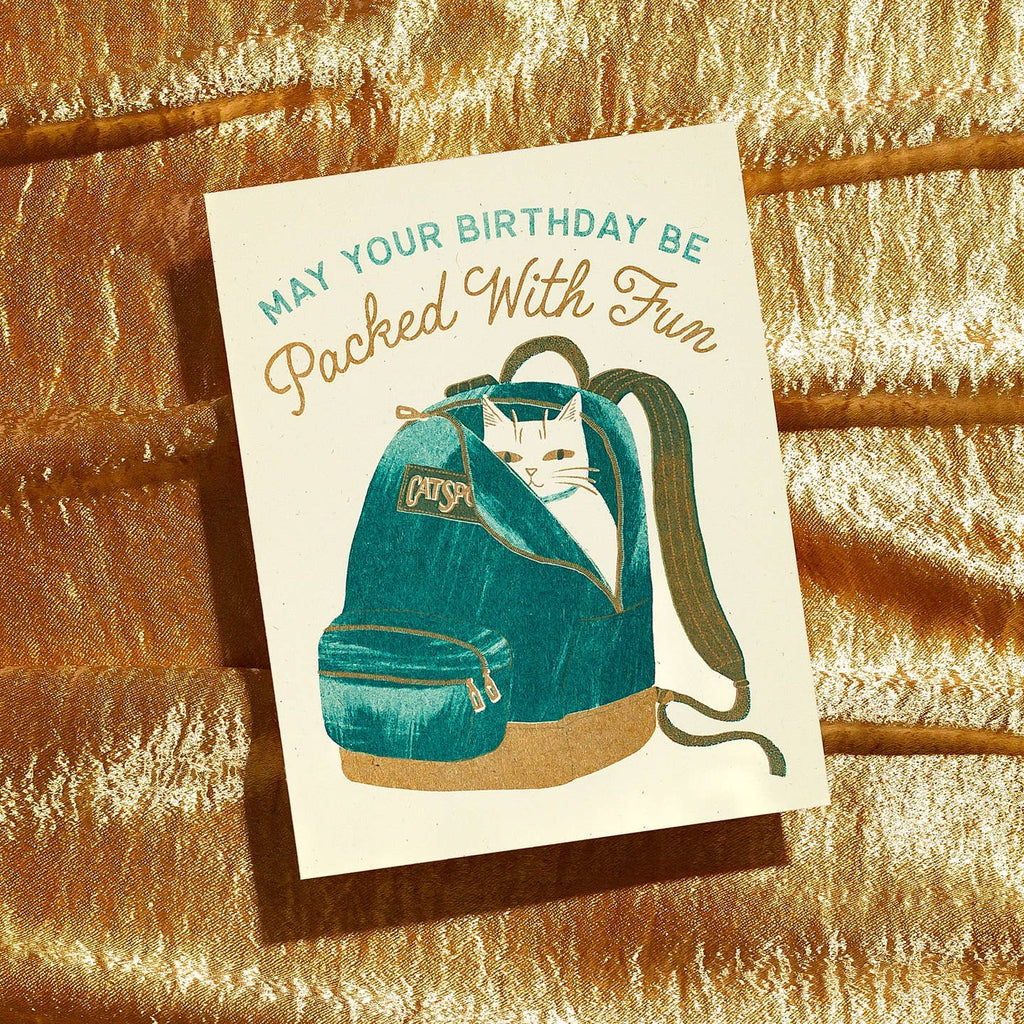 Greeting card with  cream background and image of a white cat in a teal backpack with teal text says, "May your birthday be" and tan text says, "Packed with fun". Envelope included.
