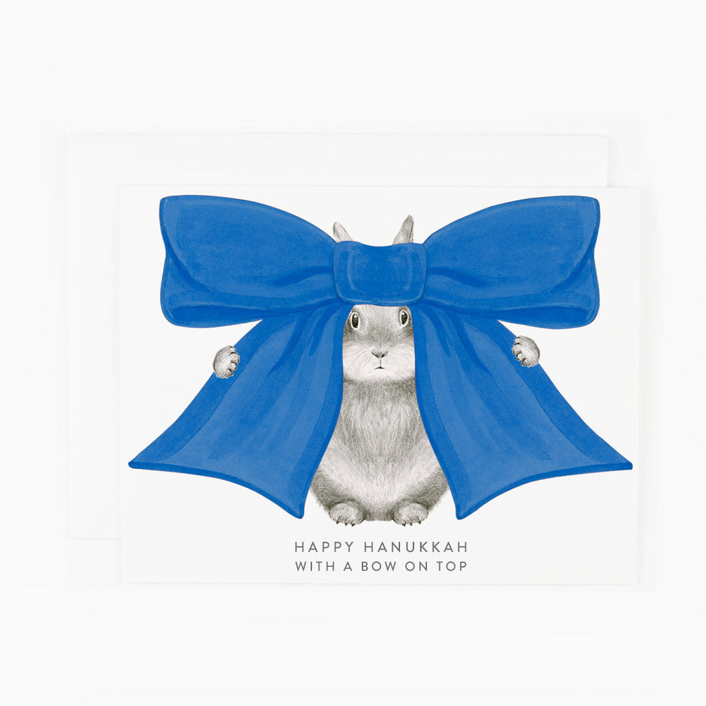 Greeting card with white background and image of a grey bunny with a big blue bow on its head. Grey text says, "Happy Hanukkah with a bow on top". Envelope included.