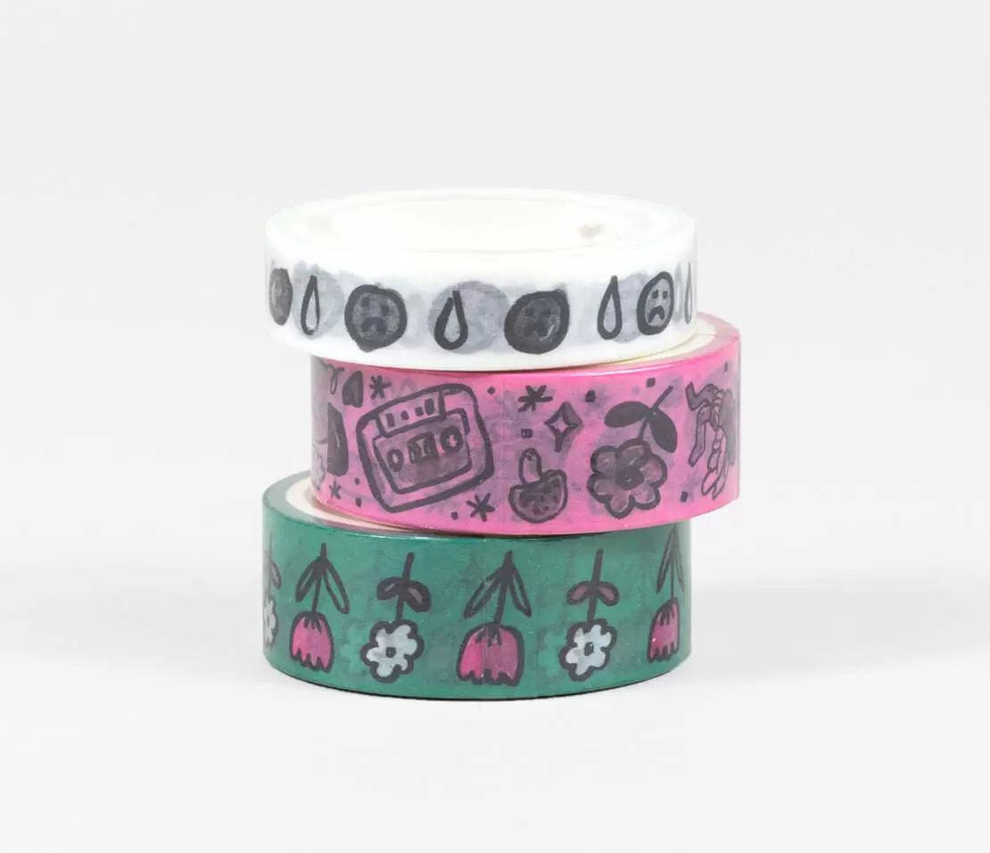 Set of three washi tapes with one with white background with grey and black sad faces and teardrops, one with pink background and doodles in black and grey of cassette tape, mushromms, flowers and one with green background with images of white daisies and pink tulips with black stems. 