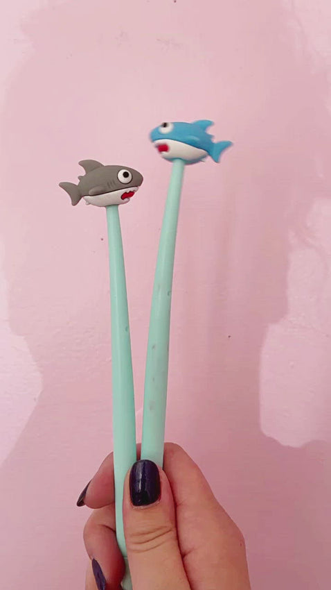 Image of four pens with pale blue barrels with sharks at the top of the pen. Bright blue shark with white underbelly and red mouth, light grey shark with white underbelly and red mouth, blue grey shark with white underbelly and red mouth and light blue shark with white underbelly with red mouth. 