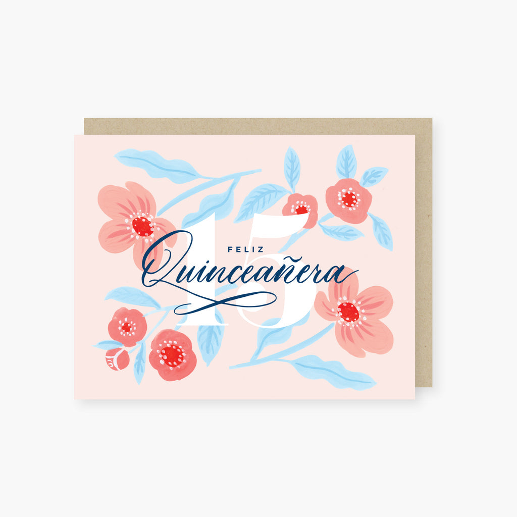 Pink card with blue text saying, “Feliz Quinceanera”. Images of pink flowers with green stems. A gray envelope is included.