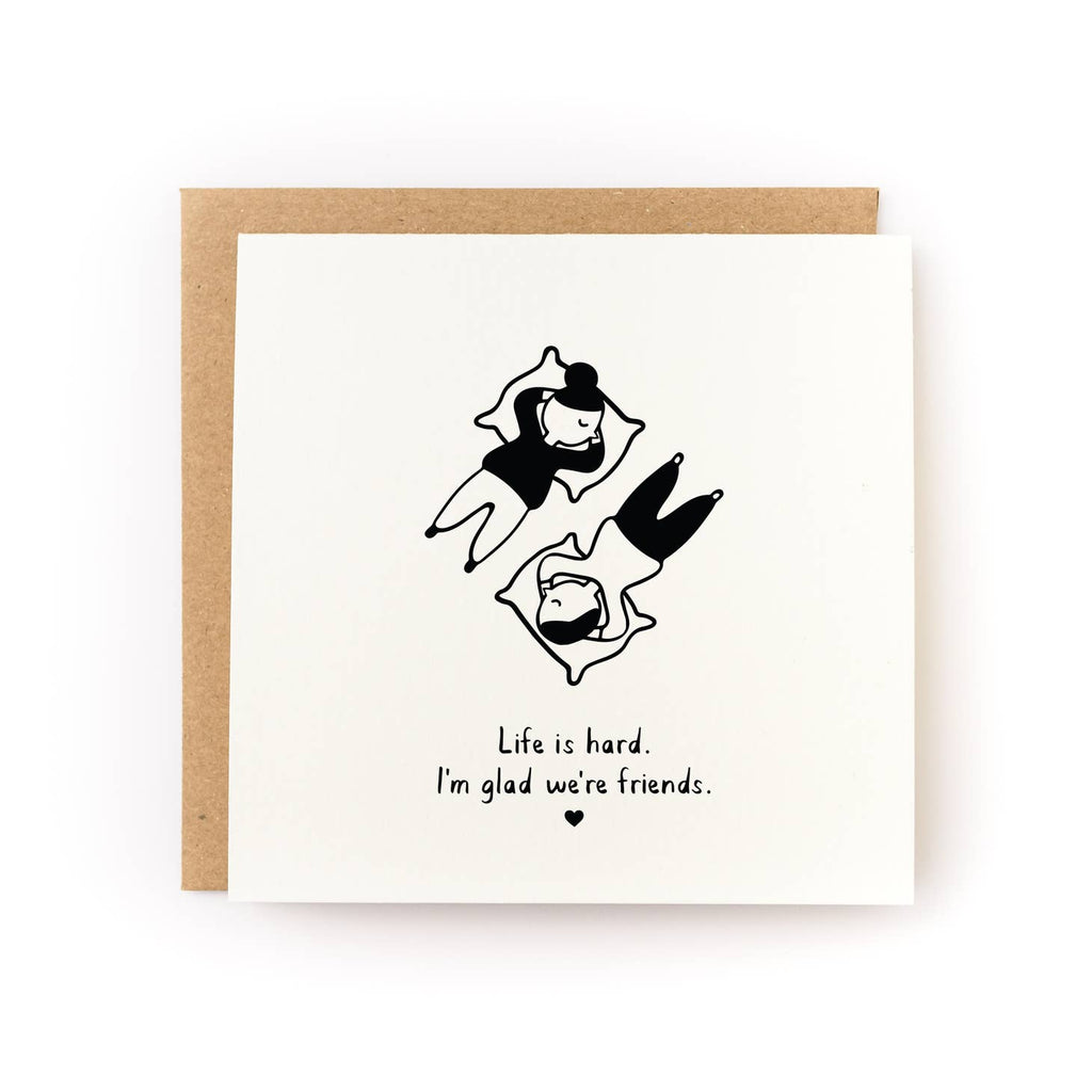 White background with two images in black and white of people lying opposite each other with pillows under their heads. Black text says “Life is Hard. I’m glad we’re friends” with a small black heart.  A brown kraft envelope is included.   