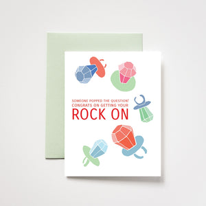 White card with red text saying, "Someone Popped the Question! Congrats on Getting Your Rock On". Images of various colored candy ring pops. A light green envelope is included.