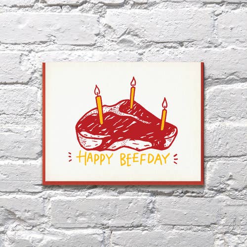 White background with image of red and white t-bone steak with three yellow  birthday candles with white flame. Yellow text says, “Happy Beefday”. A red envelope is included.   