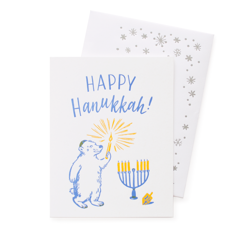 White card with blue text saying, “Happy Hanukkah!” Images of a polar bear wearing a yamika holding a lit yellow candle. Blue menorah with yellow candles and a dreidel in background. A white envelope with silver snowflakes is included.