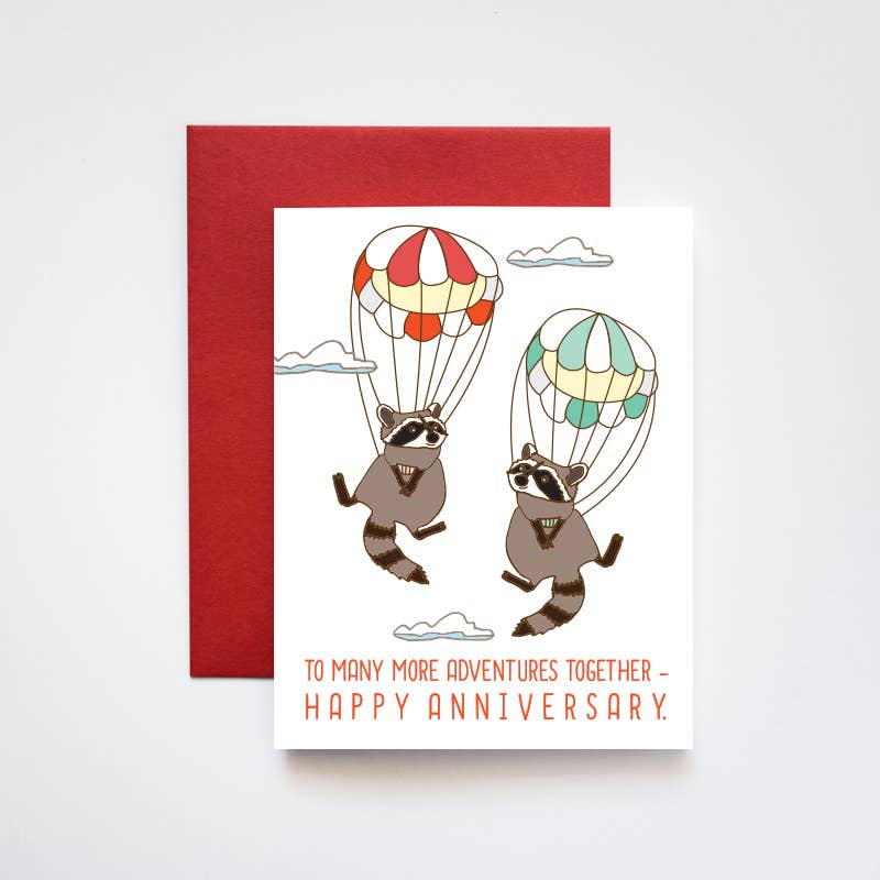 White card with red text saying, "To Many More Adventures Together- Happy Anniversary". Images of a pair of raccoons skydiving. A red envelope is included.