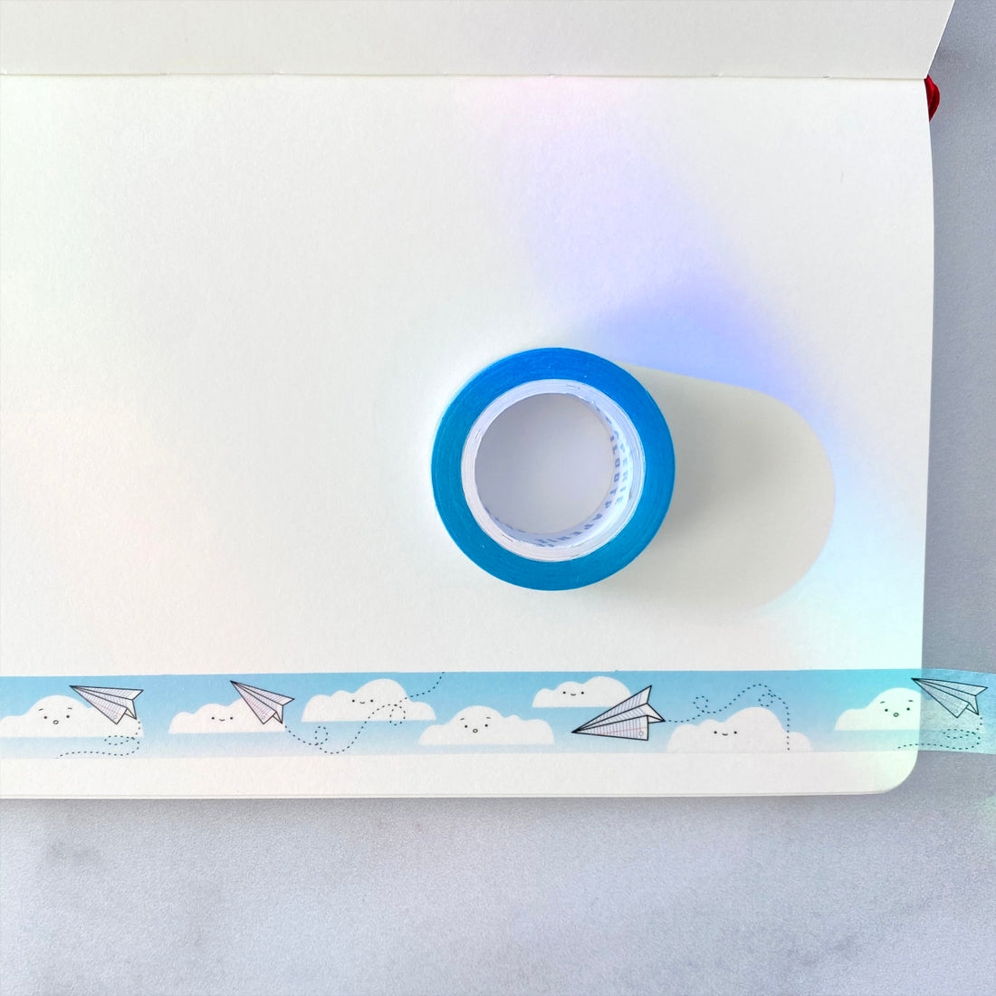 Decorative tape with blue sky background with images of white clouds and white paper airplanes zipping through the air. 