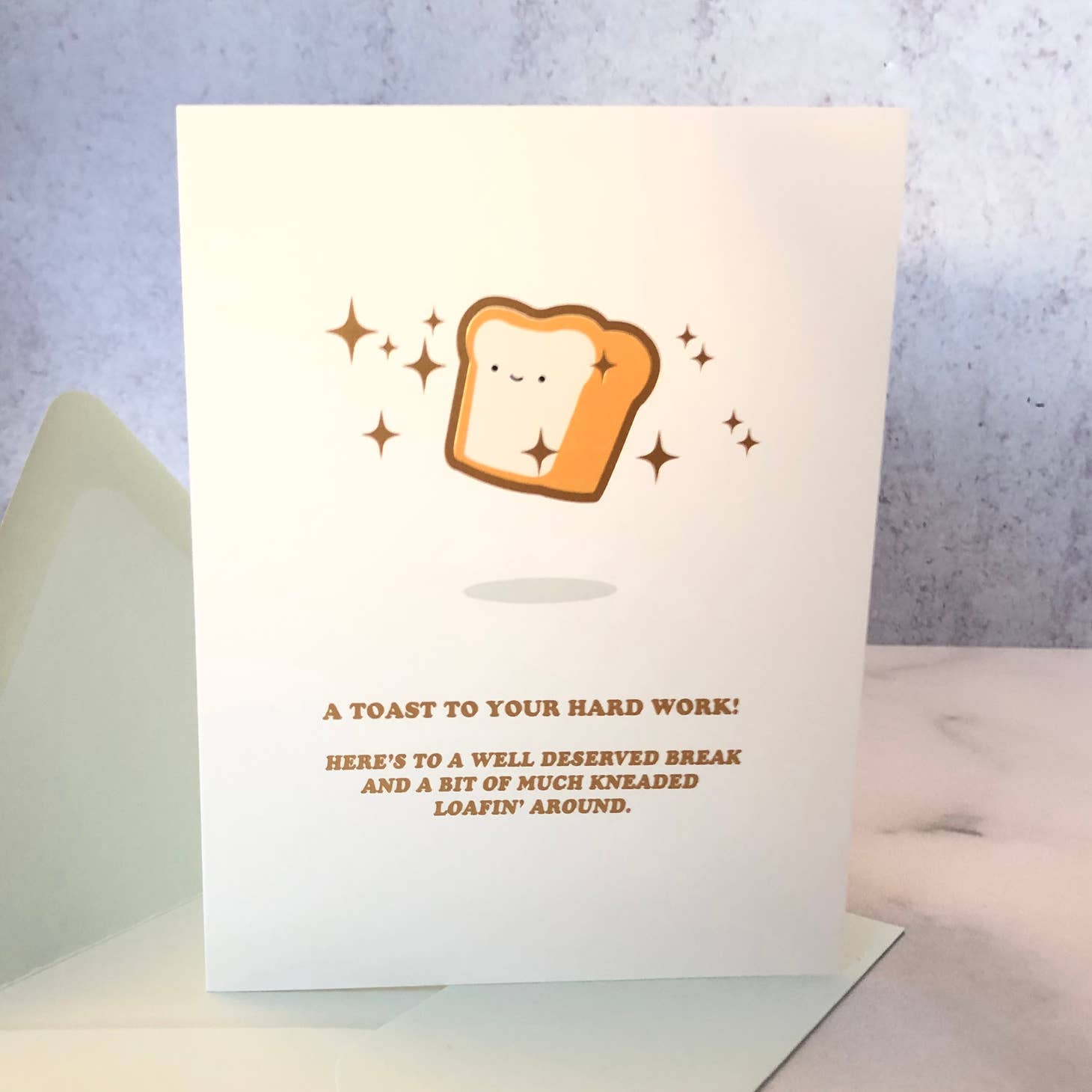 Ivory background with image of a thick piece of toast in dark brown crust with tan and off white center and black eyes and smile. Surrounded with brown sparkles.  Brown text says, “A toast to your hard work! Here’s to a well deserved break and a bit of much kneaded loafin’ around.”. An envelope is included.      