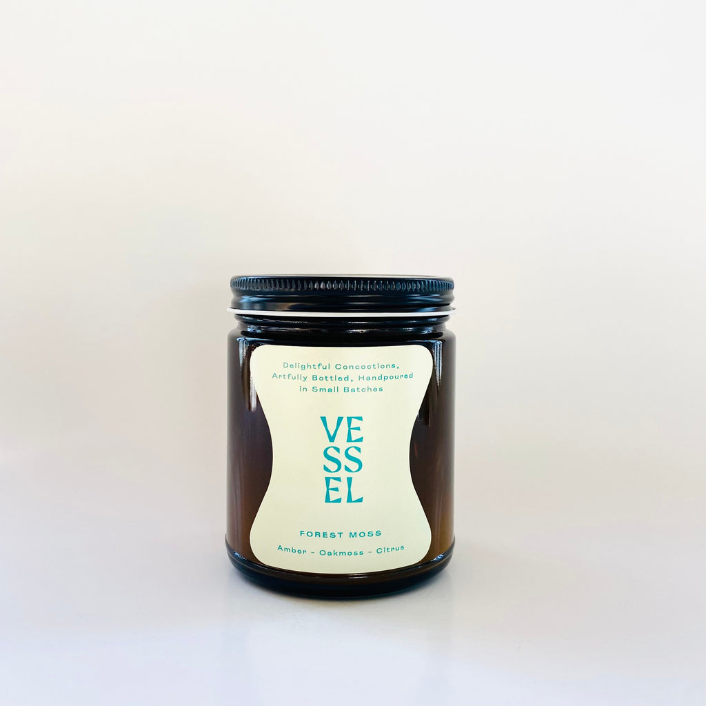 Image of brown glass jar with black lid and ivory label with teal text says, “Vessel” and “Forest Moss”. 