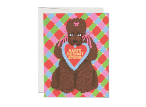 Colorful background with green, blue, purple and pink lattice. Image of a brown poodle wearing pink bows with a heart shaped card in its mouth saying, “Happy Birthday Stupid”. A white envelope is included.