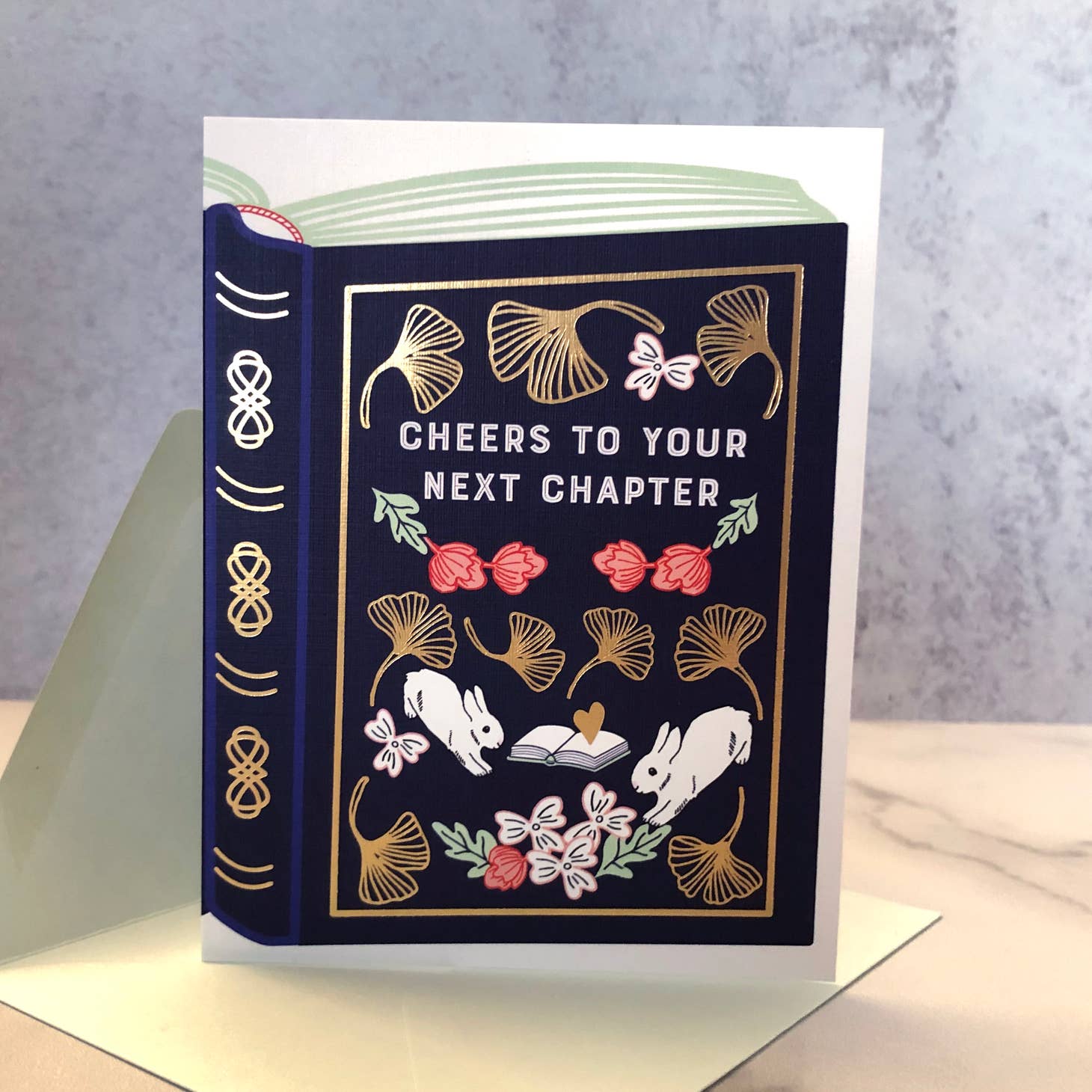 White background with large image of book in dark blue with gold foil details and grey text says, “Cheers to your next chapter” with images of pink flowers with green leaves, white rabbits, white flowers, and gold foil border. Envelope is included.    