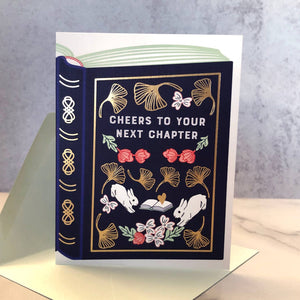 White background with large image of book in dark blue with gold foil details and grey text says, “Cheers to your next chapter” with images of pink flowers with green leaves, white rabbits, white flowers, and gold foil border. Envelope is included.    