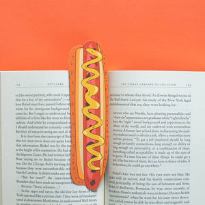Image of opened book with bookmark in the shape of a brown hotdog on a yellow bun with a drizzle of yellow mustard.