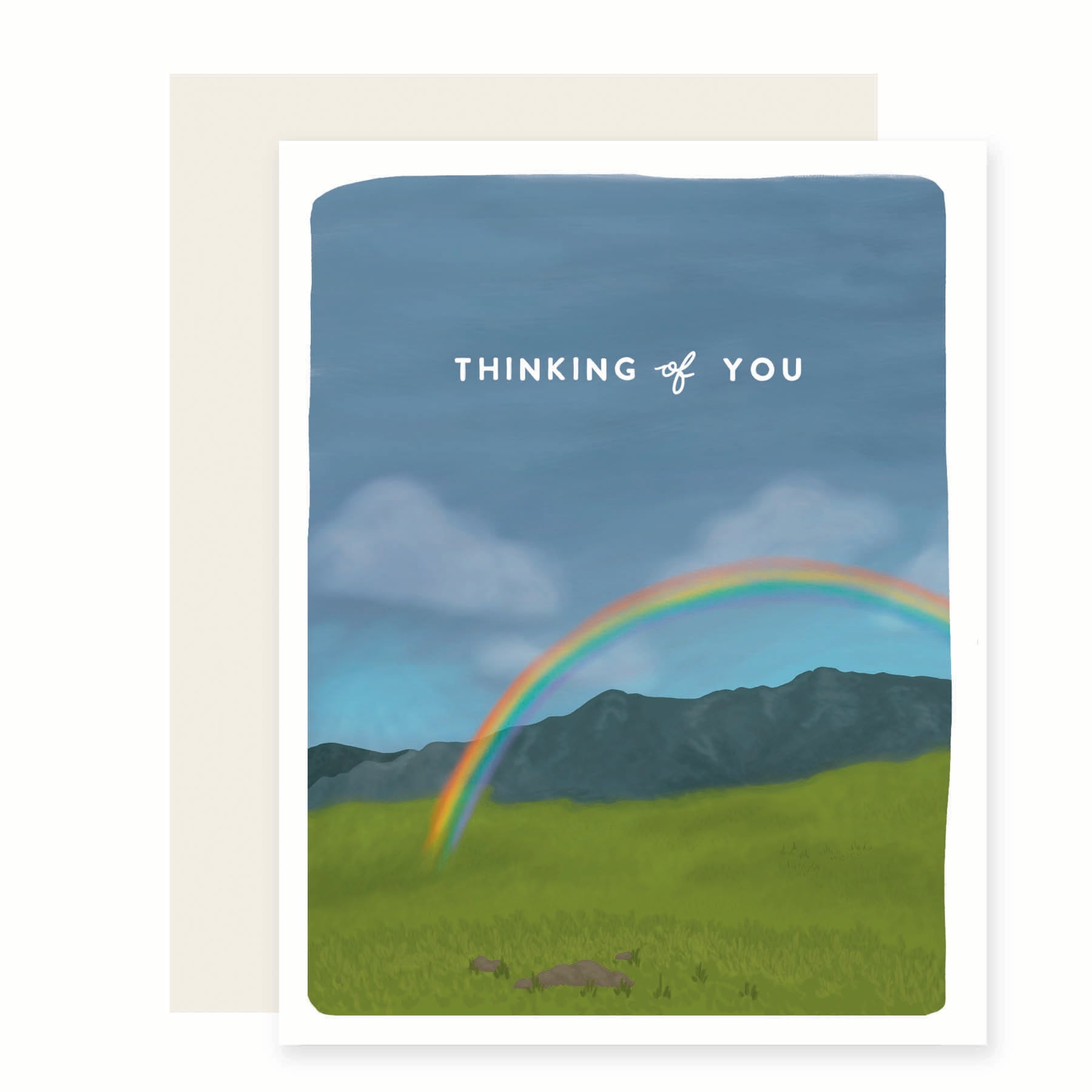 White card with meadow scene with green pasture, mountains in background, blue sky with white clouds and a rainbow across the middle of card. White text saying, “Thinking of You”. A white envelope is included.