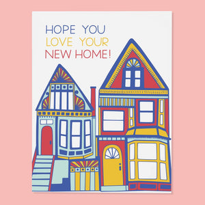 White card with blue, red and yellow text saying, “Hope You Love Your New Home!” Images of two houses next to each other. An envelope is included.