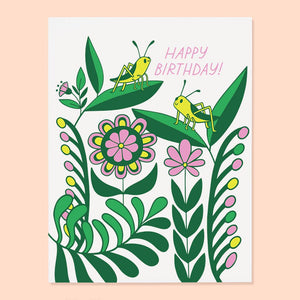 White background with image of flowers in pink, yellow and green with two yellow and green grasshoppers on the leaves with pink text says, “Happy Birthday”. Envelope is included. 