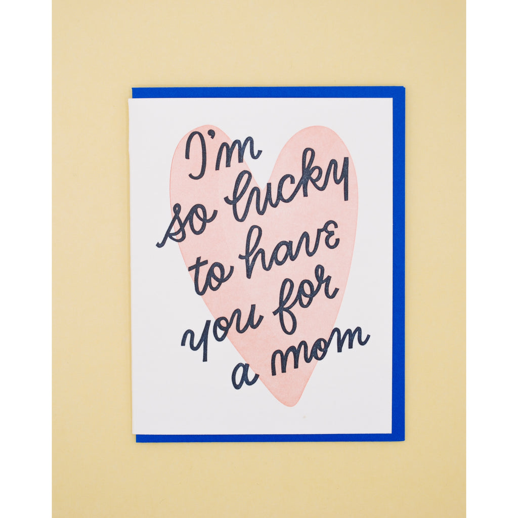 White card with black text saying, "I'm So Lucky to Have You for A Mom". Image of a light pink heart in center. A blue envelope is included.