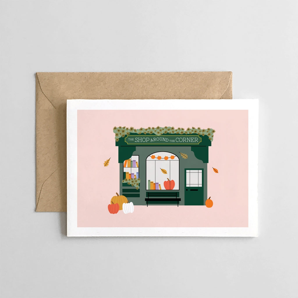 White card with peach background with an image of a green storefront with pumpkins and leaves around the doorway. Sign above store saying, "The Shop Around the Corner".  A brown envelope is included.