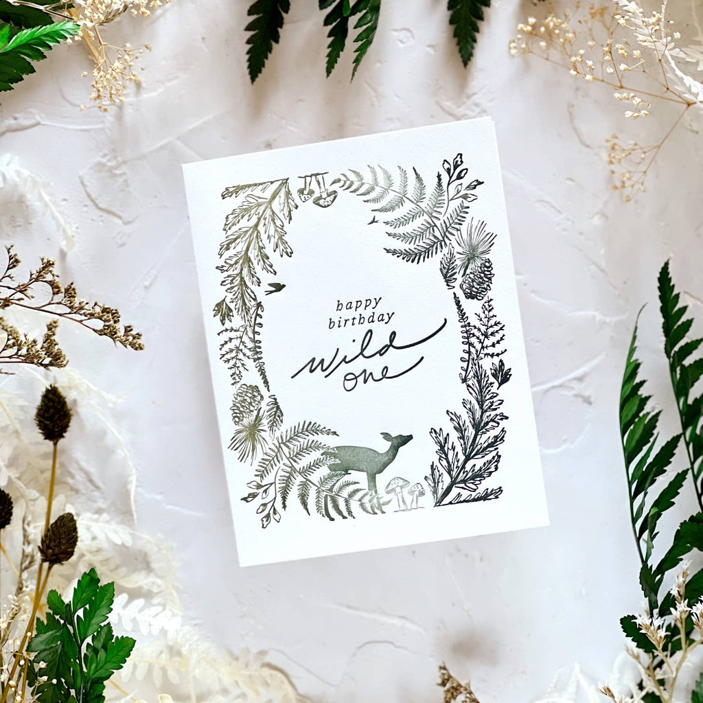 Ivory background with image of a deer and ferns, pinecones, mushrooms and pine branches in a circle around edge of card . Grey text says, “Happy Birthday Wild One”. Grey envelope included. 