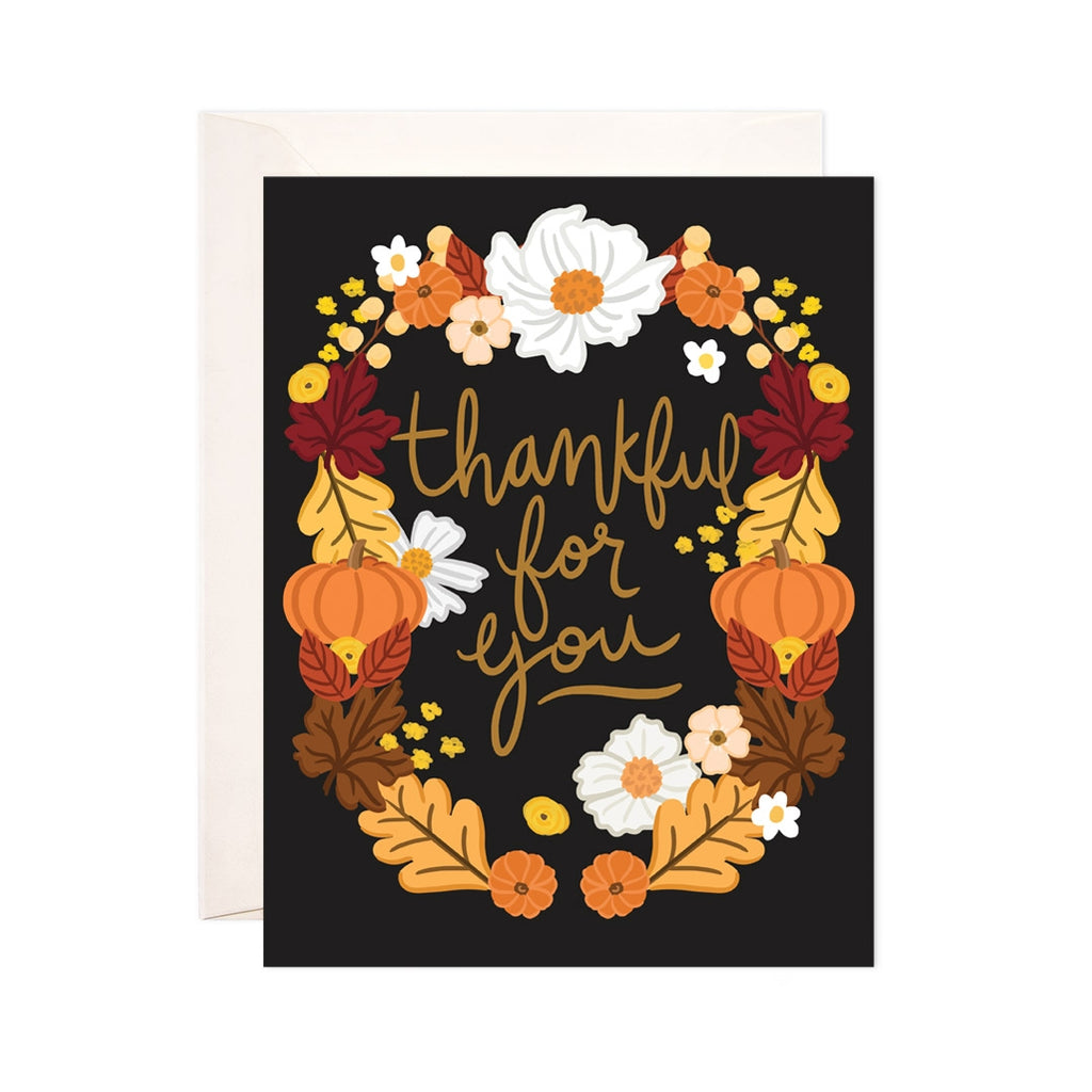 Black card with orange text saying, "Thankful For You". Image of a fall wreath with leaves and pumpkins. An ivory envelope is included.