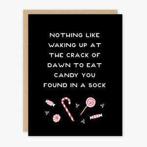 Black card with white text saying, “Nothing Like Waking Up At the Crack of Dawn to Eat Candy You Found In A Sock”. Images of candy canes, peppermint candies and holly berries. A brown envelope is included.