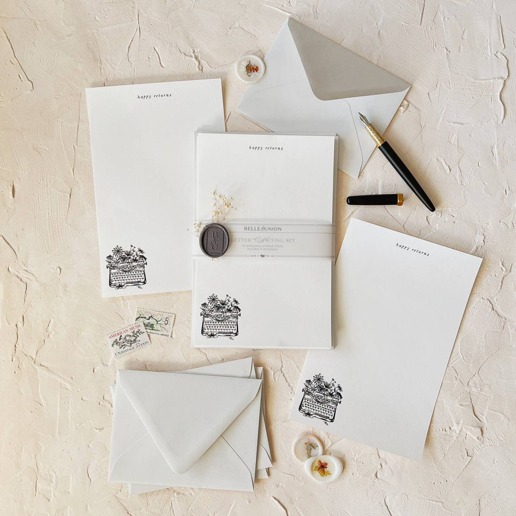 Ivory paper with image of antique typewriter adorned with wildflowers in lower left hand corner in black. Top of page has black text says, “Happy Returns”. Grey envelopes and floral seals for envelopes. 