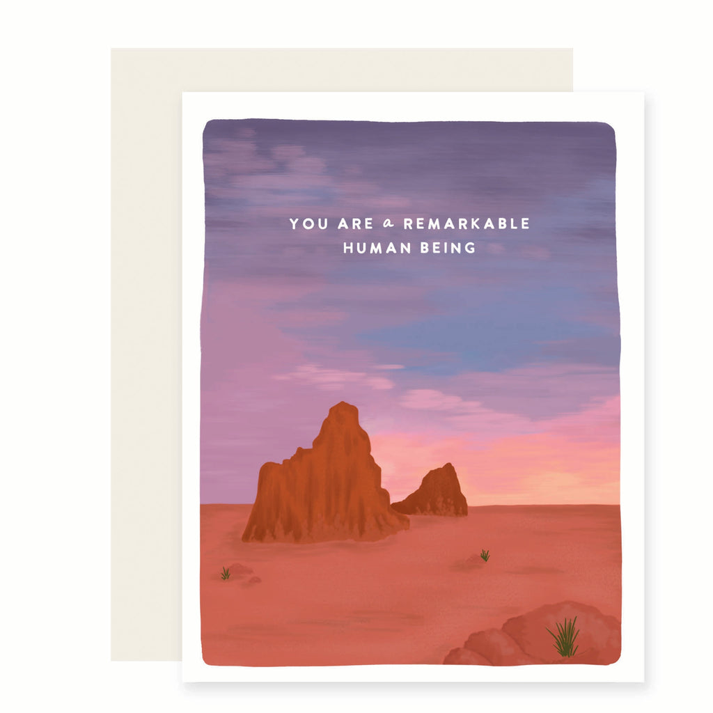 White card with desert scene including red rock formations, cactus and blue and purple sky with white text saying, “You Are A Remarkable Human Being”. A white envelope is included.