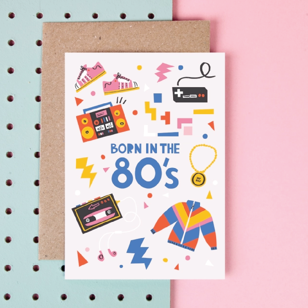 White card with blue text saying, “Born in the 80’s” Images of famous items from the 1980’s including a cassette tape, boom box, multicolored windbreaker jacket, video games, pink sneakers, and a gold necklace. Colored confetti and geometric shapes in background. A brown envelope is included.