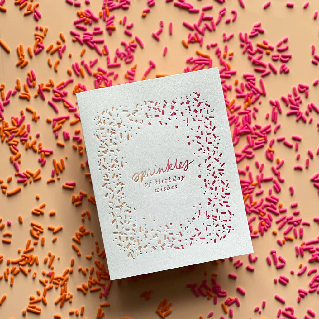 Ivory background with image of yellow, orange and pink sprinkles around edge of card. Ombre of yellow, orange and pink text says, “Sprinkles of birthday wishes”. An envelope is included. 