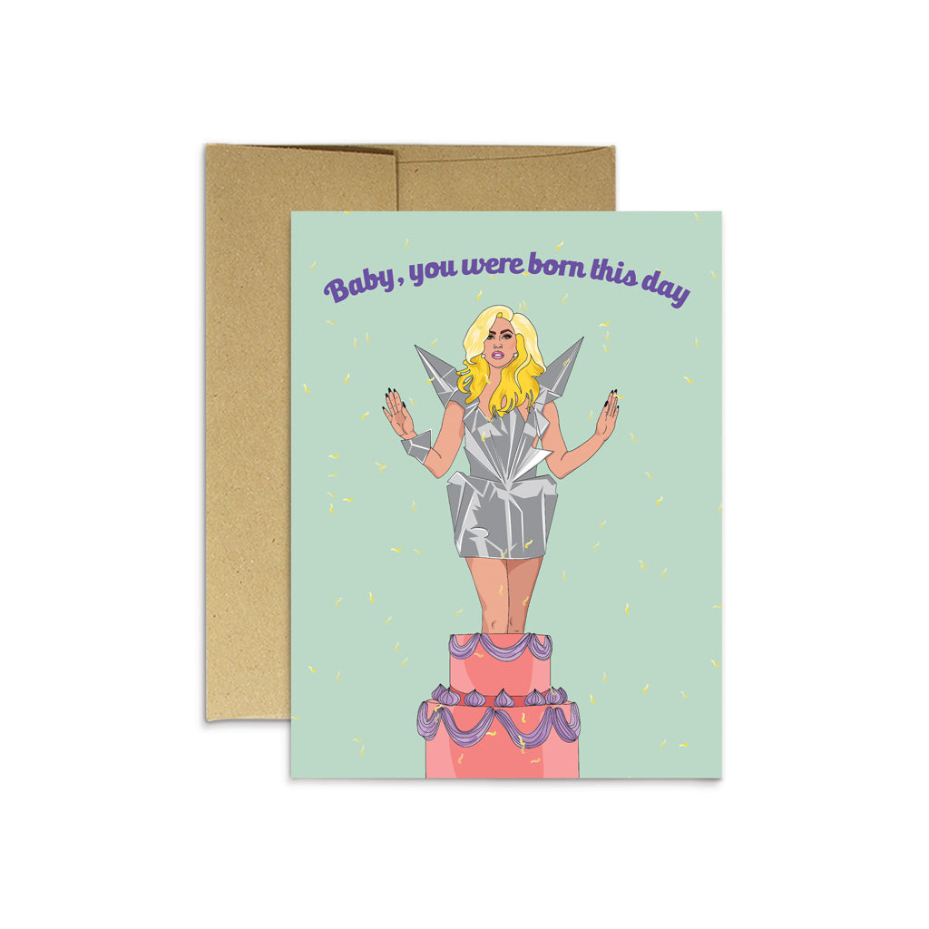 Blue card with purple text saying, “Baby, You Were Born This Day”. Image of a woman with blonde hair wearing a gray spiked dress coming out of a 2 tier pink and purple birthday cake. A brown envelope is included.