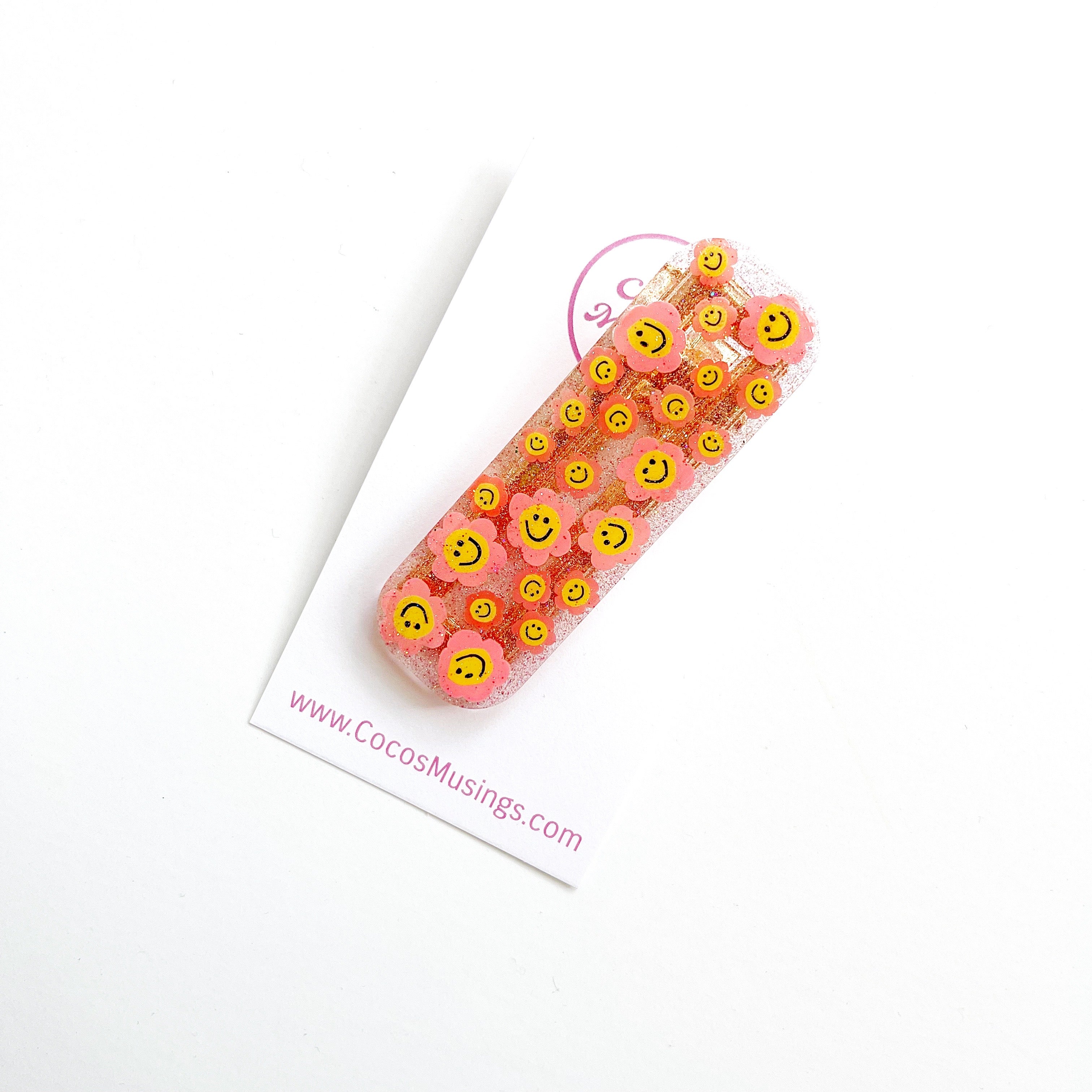 Image of clear clip with glitter and images of pink flowers with yellow smiley faces in the center on a rectangular hair clip.  