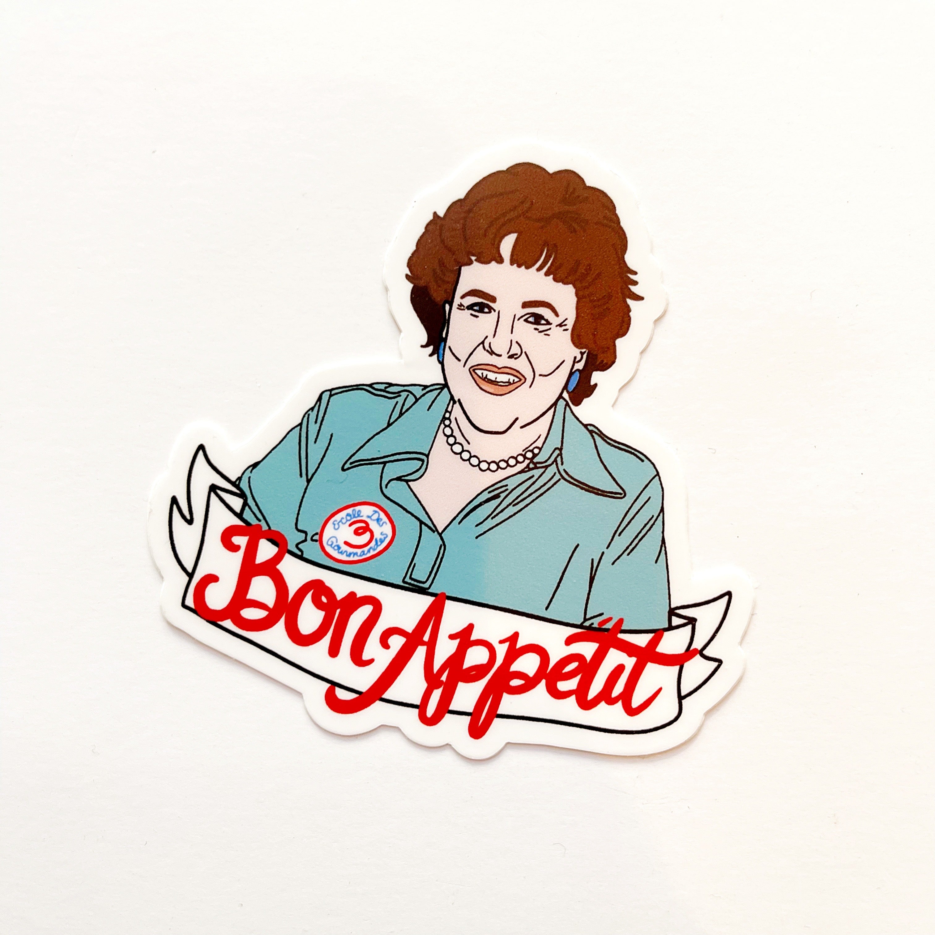 Image of Julia Child in a blue shirt with red text on a white banner says, “Bon Appetit”.