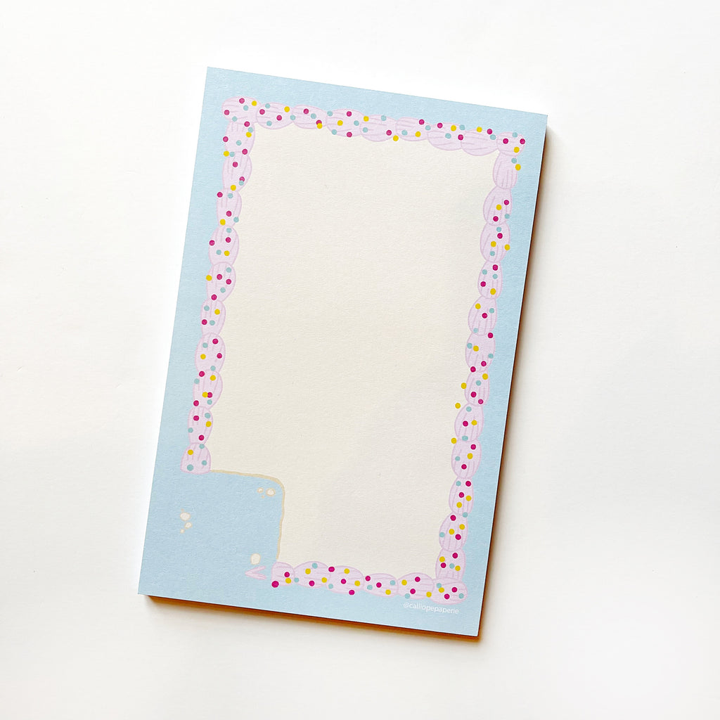 Image of notepad with white background bordered with light blue and pink frosting rectangle with colorful sprinkles with a square corner removed. 