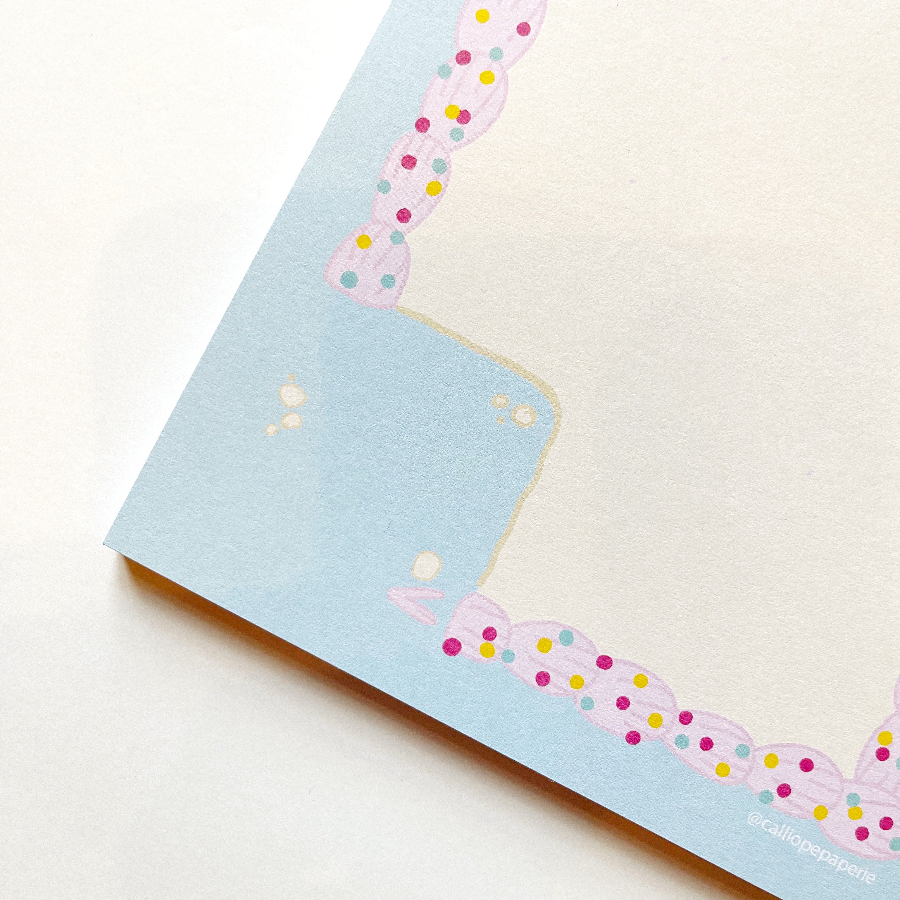 Image of notepad with white background bordered with light blue and pink frosting rectangle with colorful sprinkles with a square corner removed.