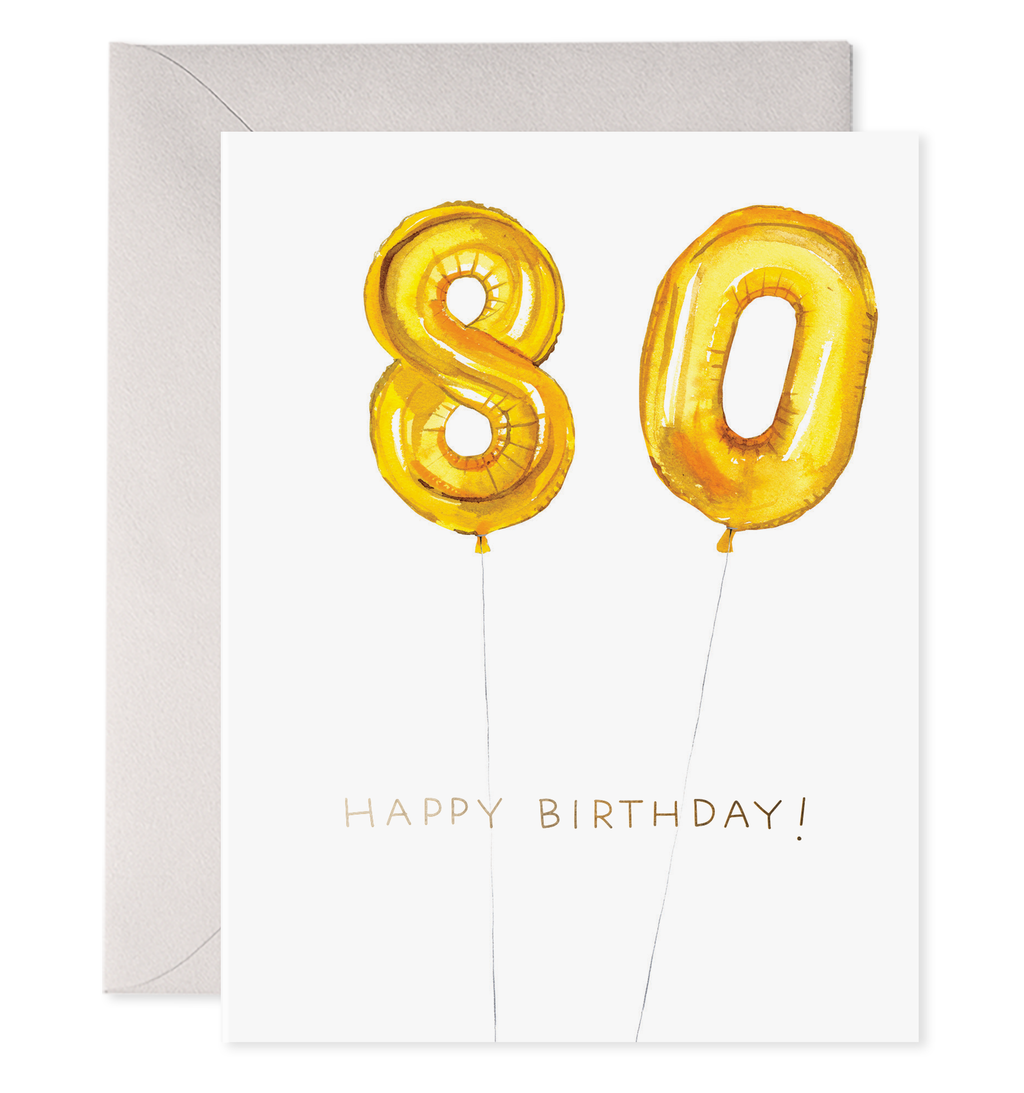 White card with black text saying, “Happy Birthday”.  Images of gold foil balloons in the shape of an eight and a zero. A purple envelope is included.