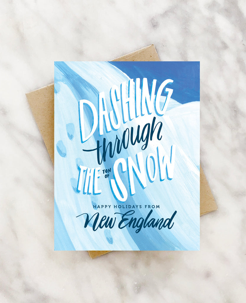 White card with white and blue text saying “Dashing through the Ton of Snow Happy Holidays from New England”. Images of white snowhills. A brown envelope is included.