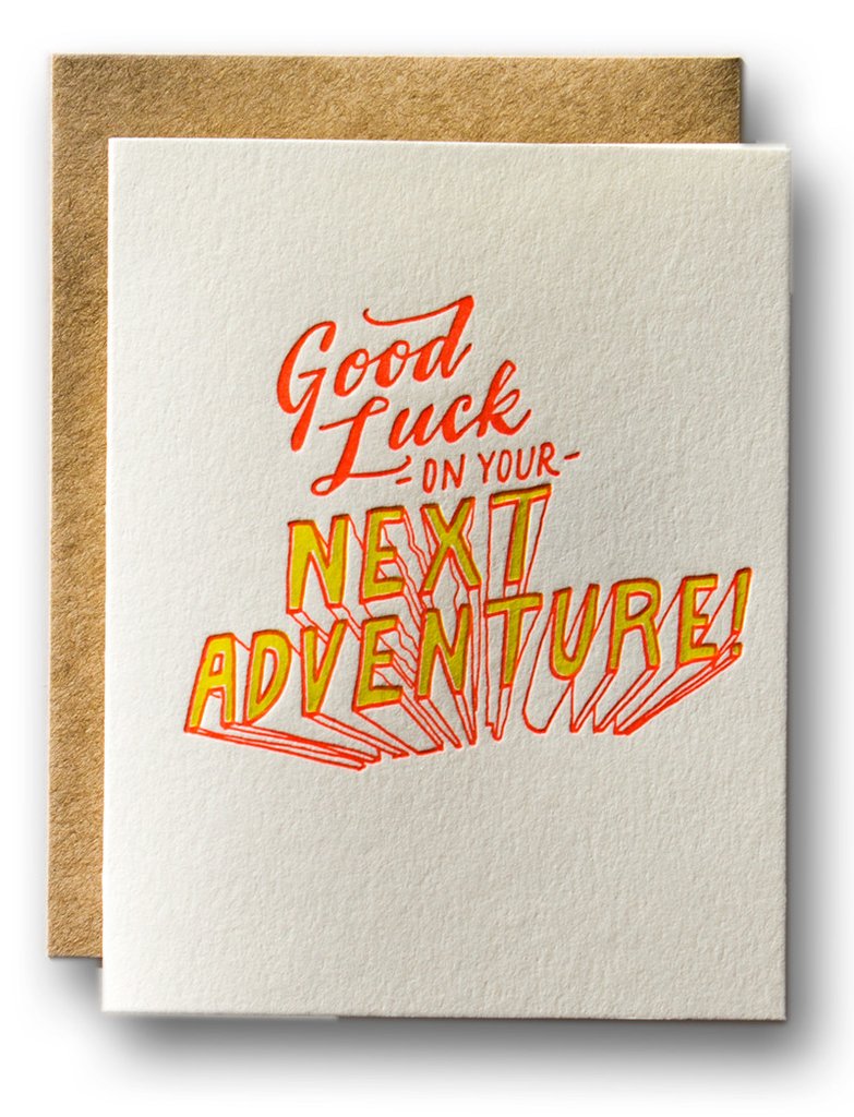 Gray card with orange and yellow text saying, "Good Luck on Your Next Adventure". Image of Next Adventure text is yellow with 3D lines extending the letters. A brown envelope is included.