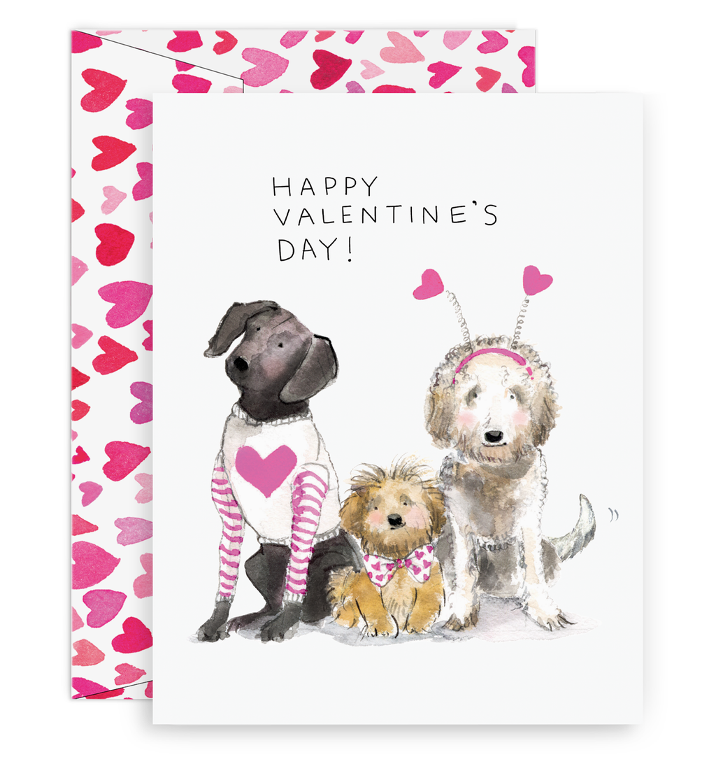 White background with image of three dogs. Black dog in a white shirt with a pink heart on the front and pink and white striped sleeves, small tan dog wearing a white bowtie with red hearts. White dog with red headband with two red heart antennas. Black text says, “Happy Valentine’s Day”. A white envelope with red and pink hearts is included.   