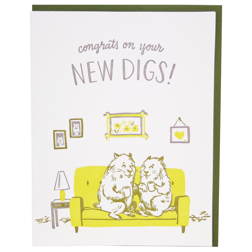 White card with gray text saying, “Congrats On Your New Digs!” Images of two groundhogs sitting on a yellow couch in a living room with pictures hung on the wall and table with lamp. A green envelope is included.