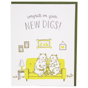 White card with gray text saying, “Congrats On Your New Digs!” Images of two groundhogs sitting on a yellow couch in a living room with pictures hung on the wall and table with lamp. A green envelope is included.