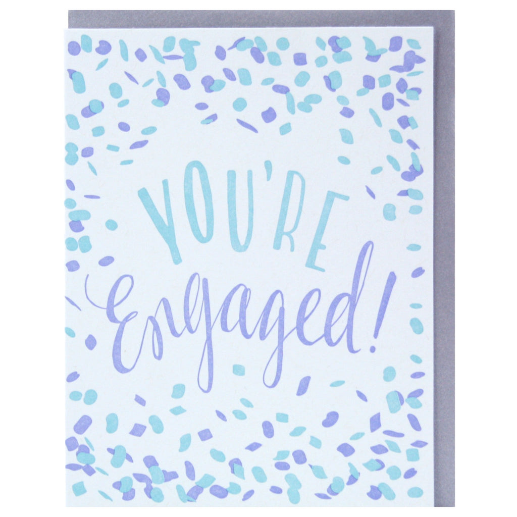 Light blue card with blue and purple text saying, “You’re Engaged!” Images of blue and purple confetti scattered across card. A purple envelope is included.