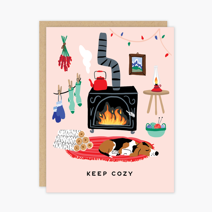 Pink card with black text saying, “Keep Cozy”. Images of a living room scene with a black stove with a fire, a brown dog laying on a red rug, white logs for the fire, socks hanging on a clothesline, and Christmas lights strung along the ceiling. A brown envelope is included.