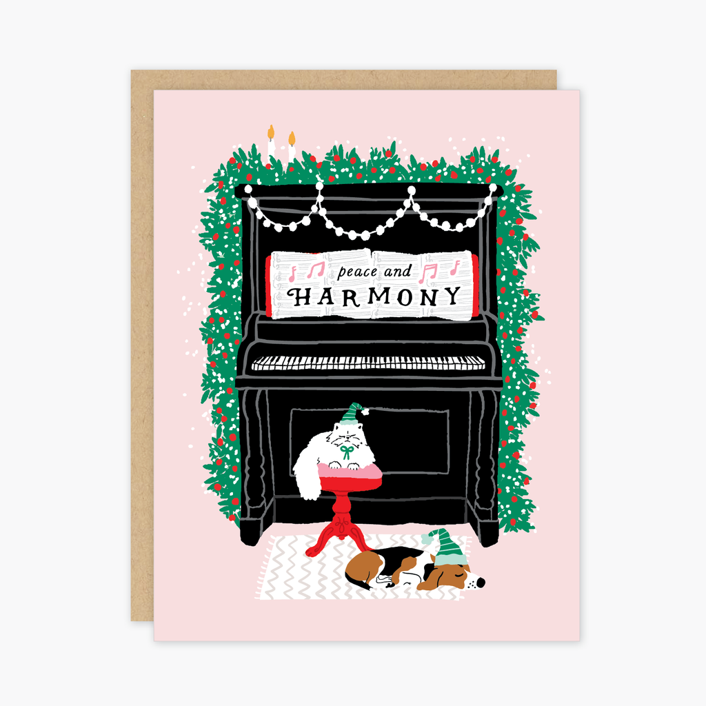 Pink card with images of a large black piano decorated with green garland and white garland with candles on top. A white cat and brown dog in front of piano. White sheet music on piano with black text saying, “Peace and Harmony”. A brown envelope is included.