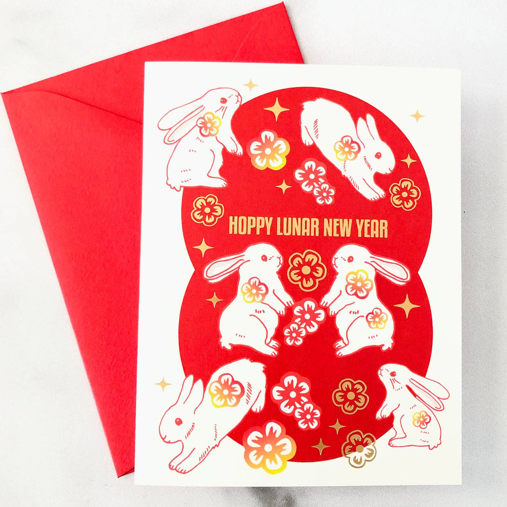 White card with gold foil text saying, “Hoppy Lunar New Year”. Images of several rabbits hopping across card with gold foil flowers. A red envelope is included.