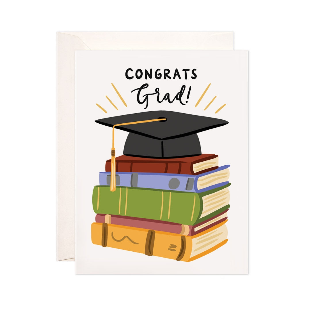 Ivory card with black text saying, "Congrats Grad!". Image of a stack of books with a black graduation hat on top. An ivory envelope is included.