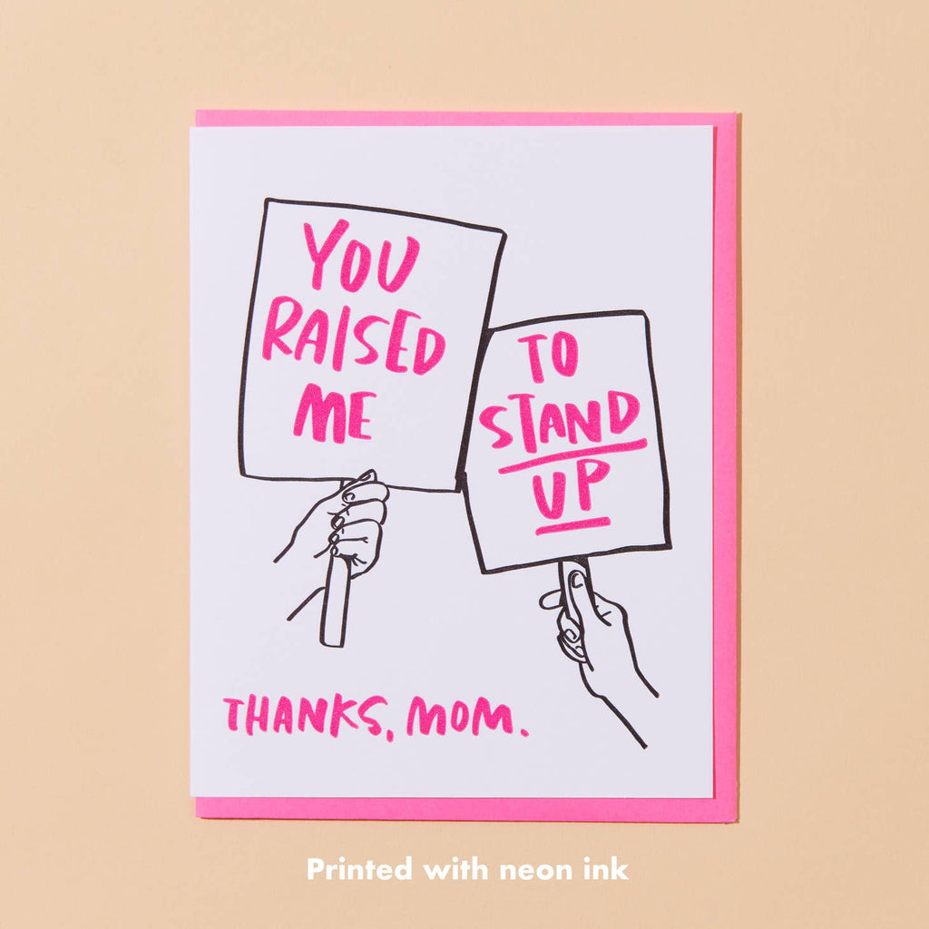 White card with two hands holding posters, one says “You raised me” in hot pink text and the other says “to stand up” in hot pink text. “Thanks, Mom.” in hot pink text at bottom of card. Hot pink envelope included.        