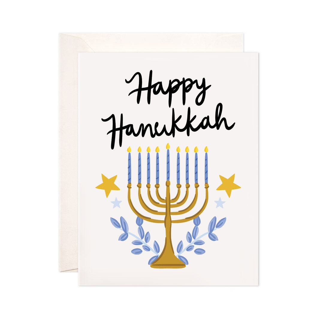 Ivory card with black text saying, "Happy Hanukkah". Image of a gold menorah with blue branches and gold stars. An ivory envelope is included.